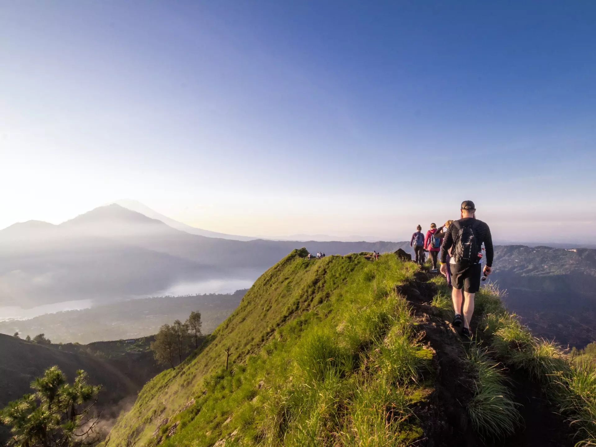 Hikers walk along the ridge of a mountain. Silhouettes of other mountains are visible through mist in the distance.