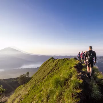 Hikers walk along the ridge of a mountain. Silhouettes of other mountains are visible through mist in the distance.