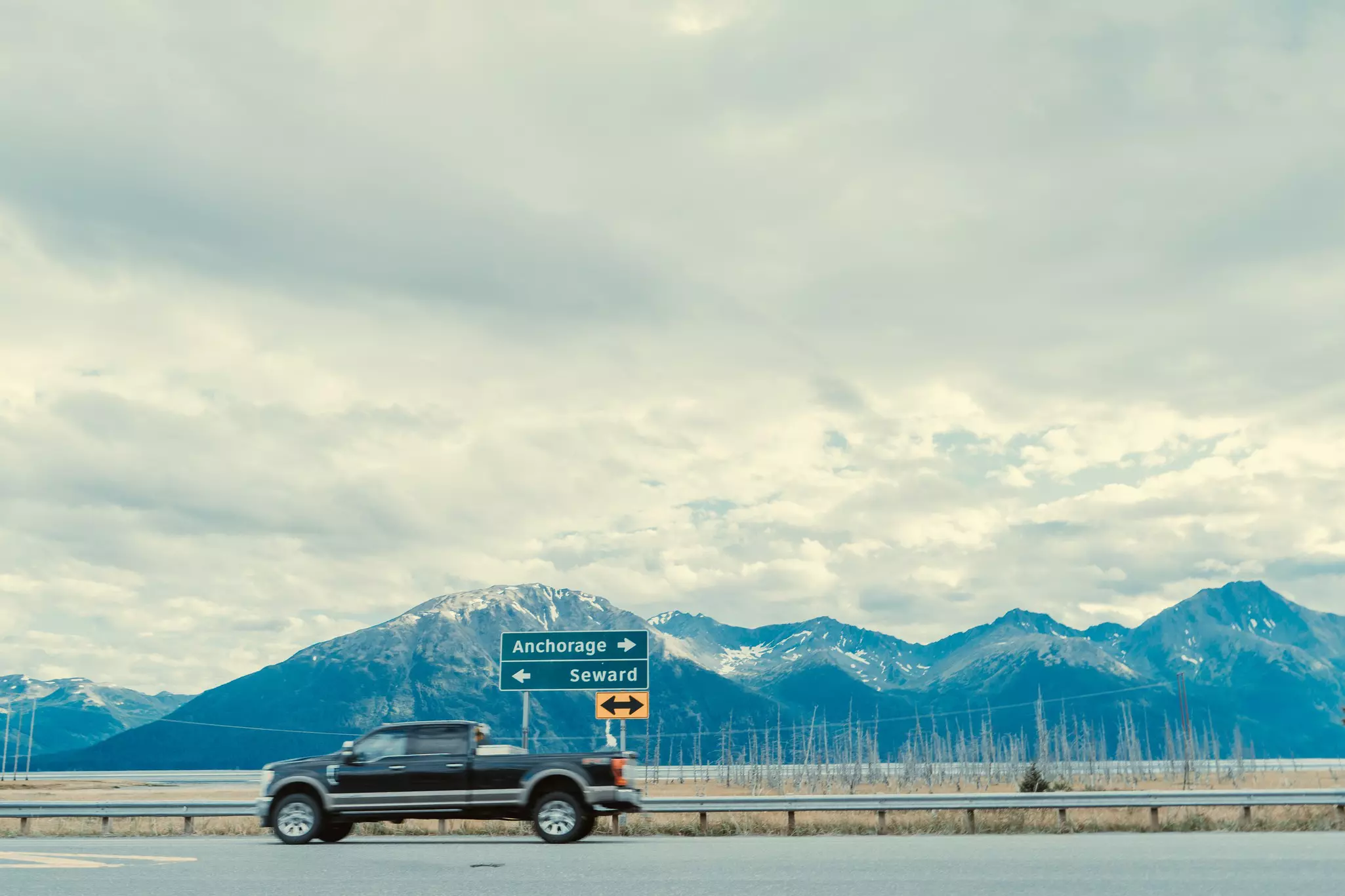 A pickup truck drives on a highway past snow-covered mountains.