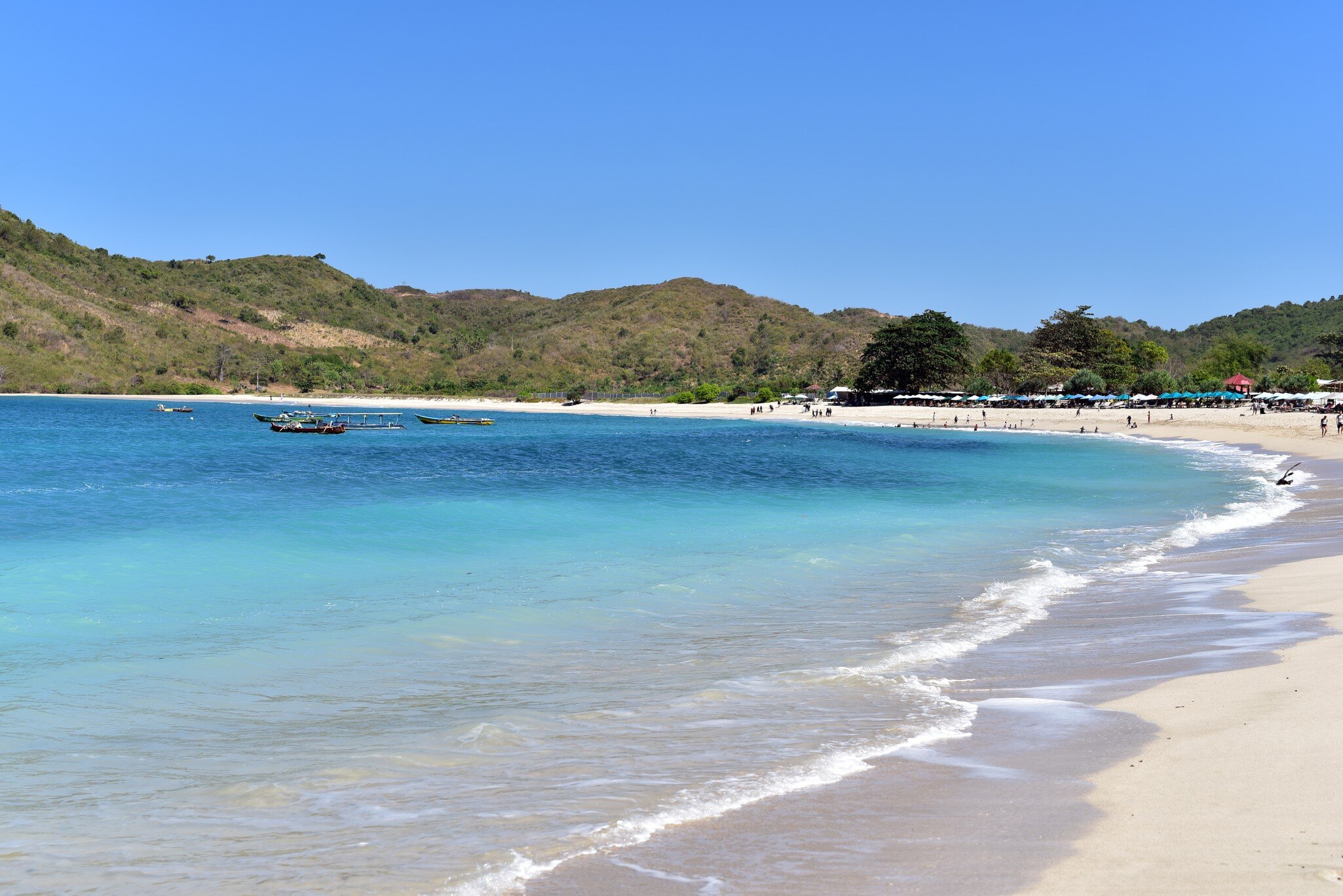Turquoise water laps a pale sand beach backed by green hills, with outrigger boats and beach umbrellas