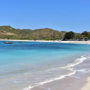 Turquoise water laps a pale sand beach backed by green hills, with outrigger boats and beach umbrellas