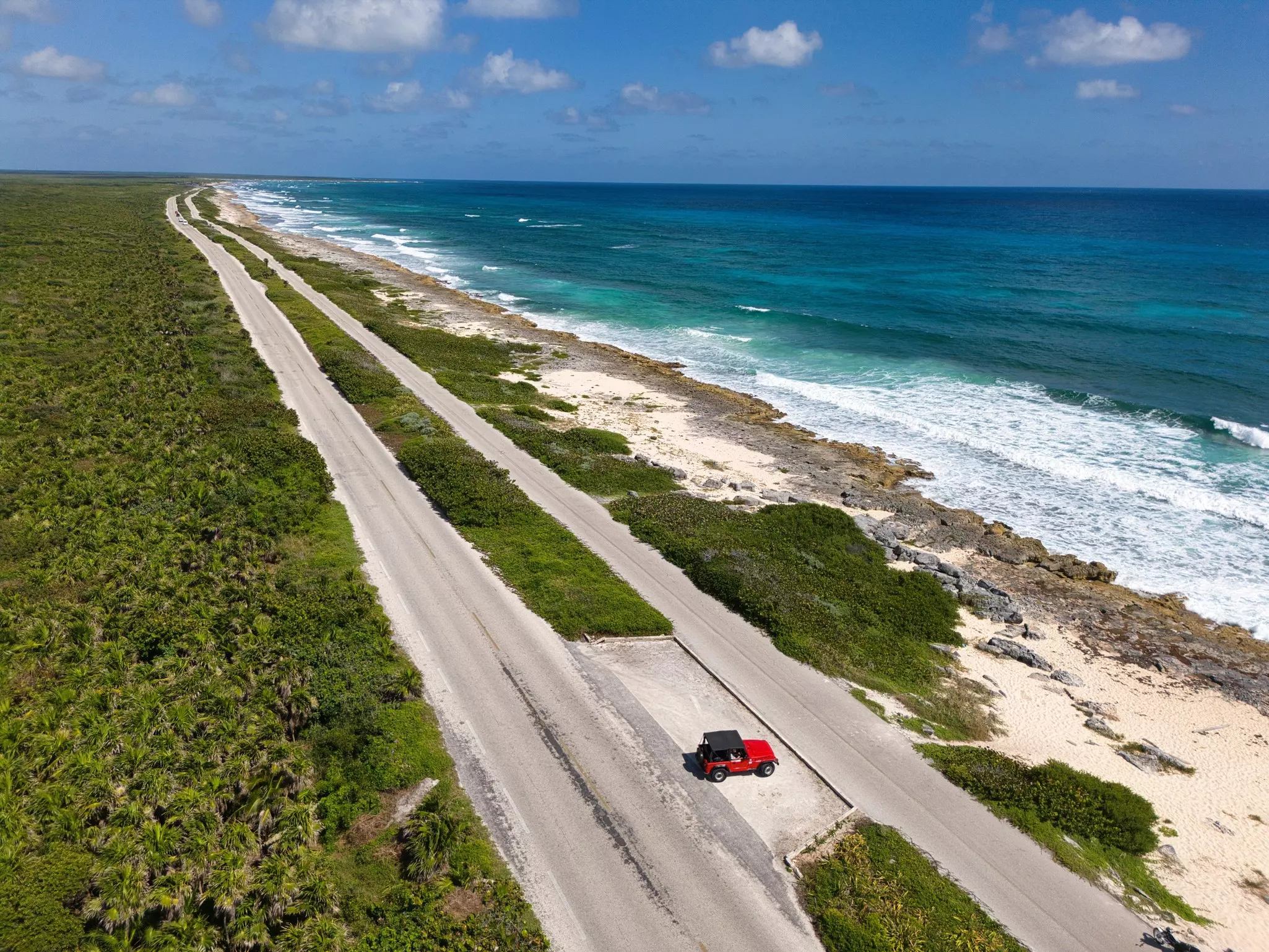 An aerial view of a red Jeep parked next to a deserted road by a tropical beach.