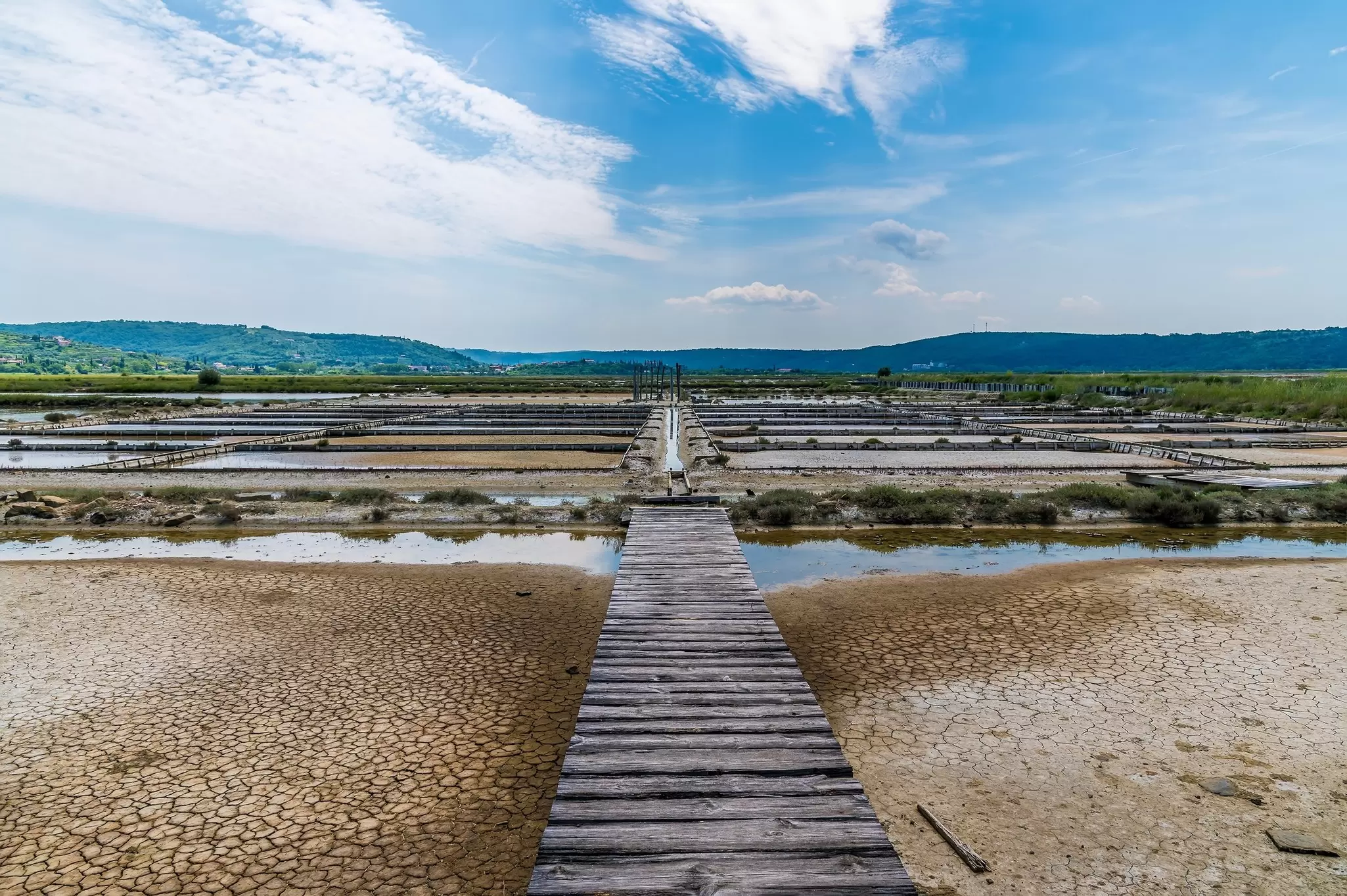 Wooden boardwalk leading across cracked mud between the grid of shallow evaporation basins of a coastal saltworks under a blue sky