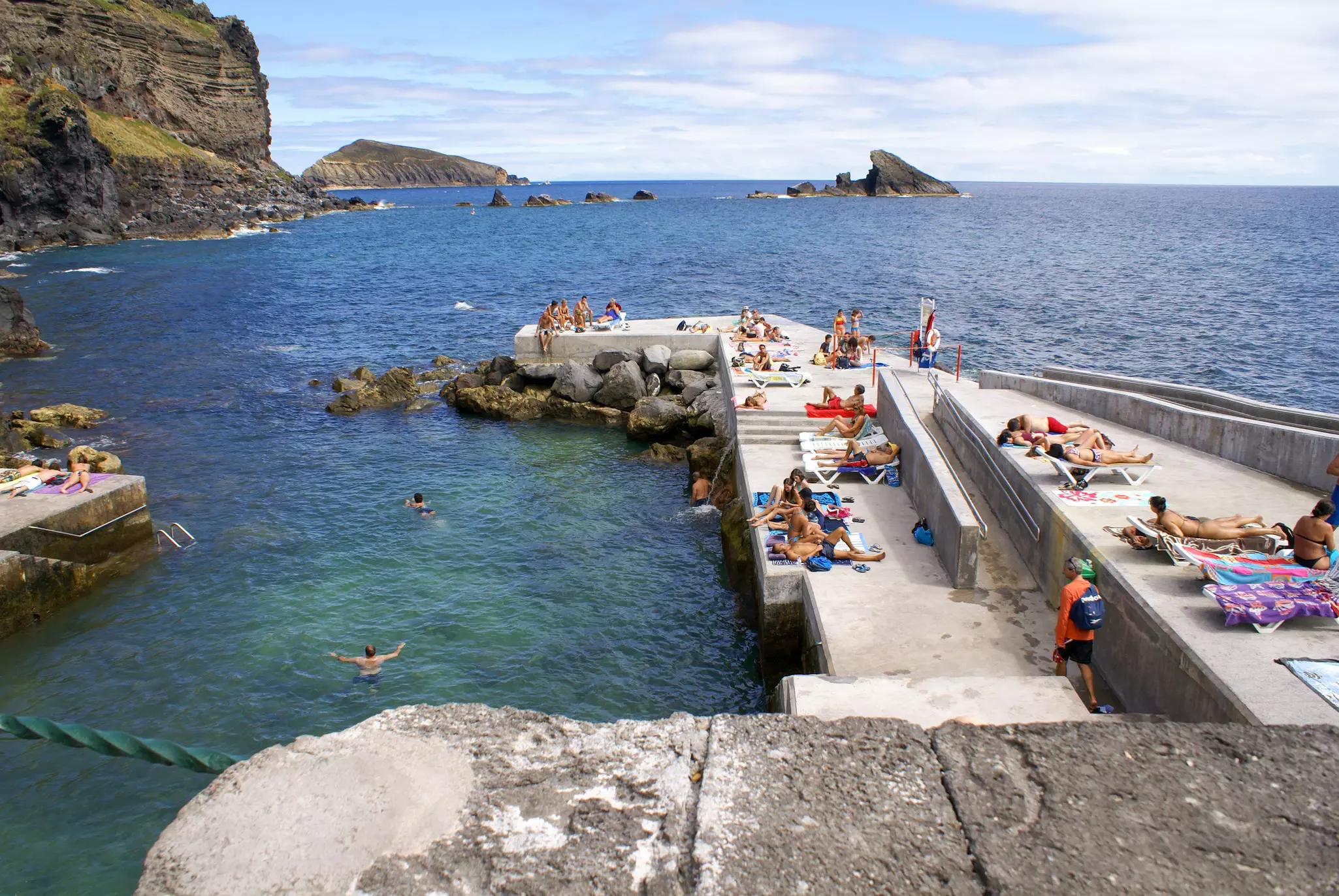 People lie out in the sun on a concrete pier built atop rocks jutting into the sea. Some people swim in the water.