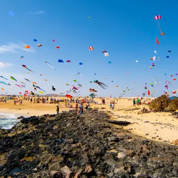 Hundreds of colorful kites fly over a sandy beach on a windy day; there is a rocky outcrop in the foreground and people on the beach under the kites.