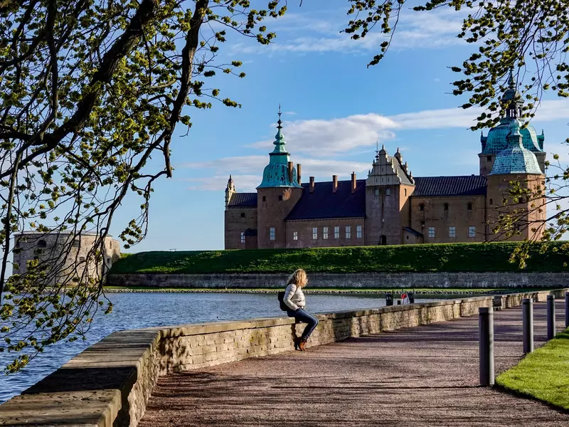 A woman sits on a stone wall by a path along the water; there is a castle in the background.
