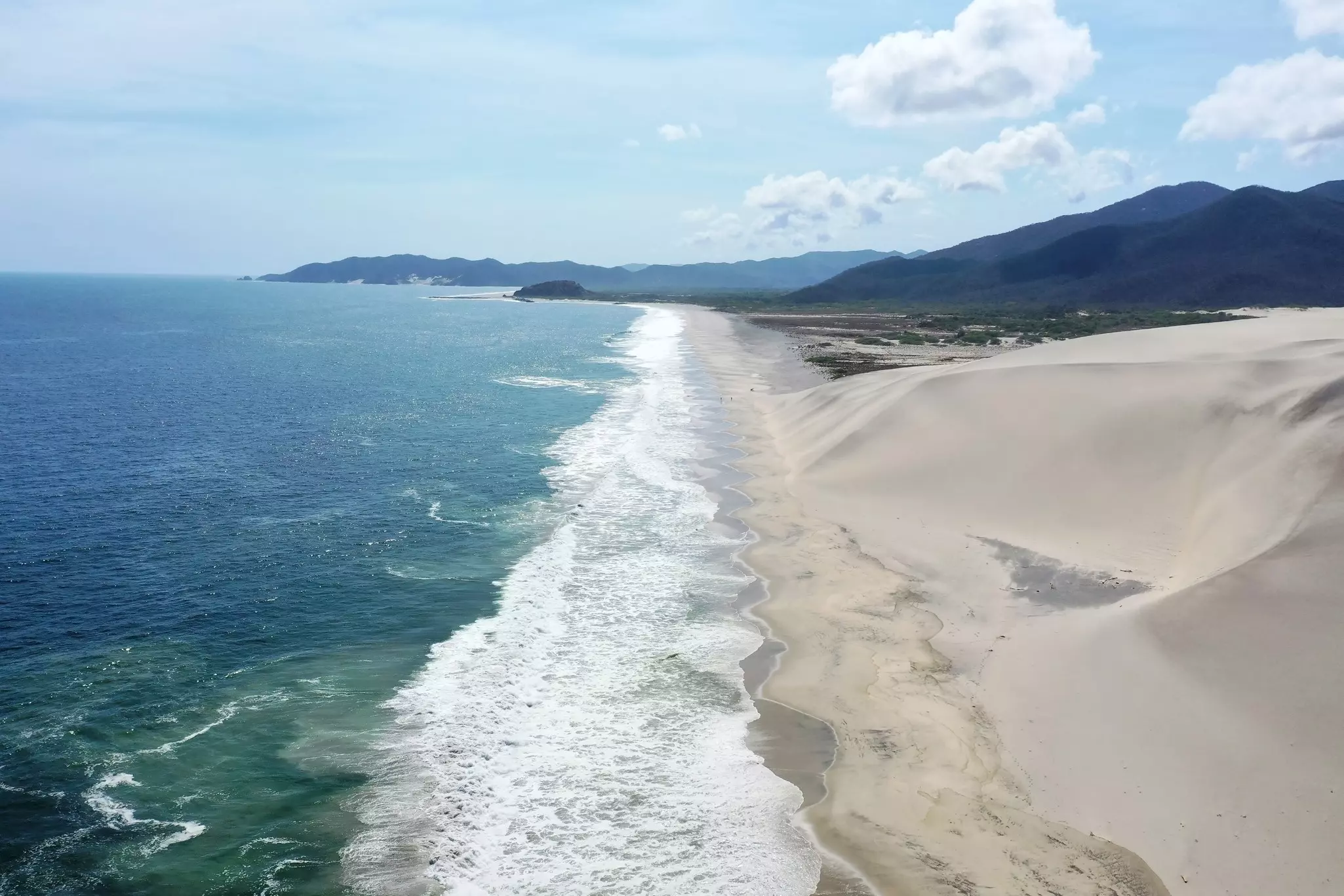 A deserted beach with foaming waves and large dunes on shore.