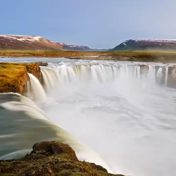 Wide horseshoe waterfall pouring into a misty gorge, mossy rocks in front and snow-patched hills under a pale sky.