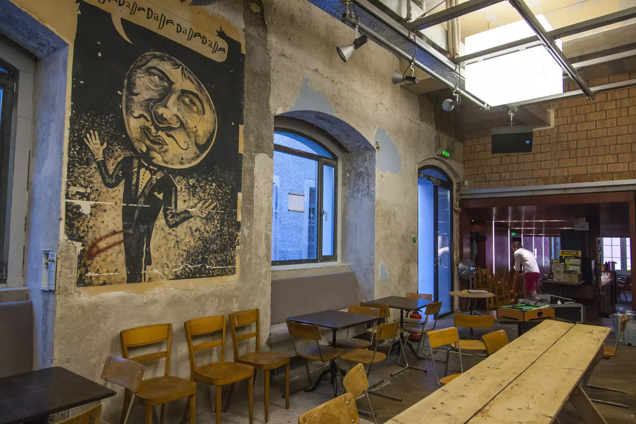 A meeting space has a narrow table and chairs. A mural showing a caricature of a man is on one wall.