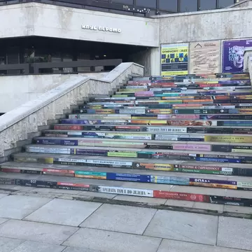 Wide stone staircase whose risers are printed with colourful book titles, concert posters beside a concrete building.