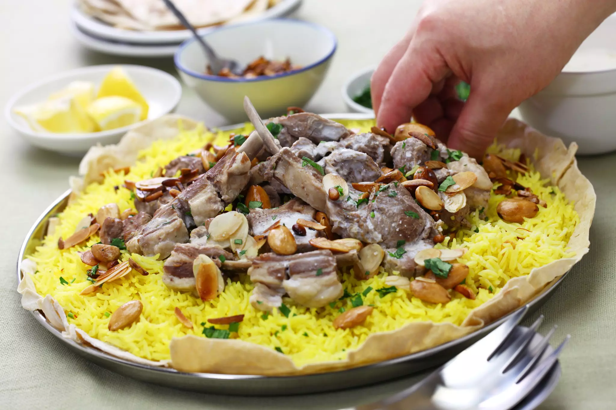 A hand reaches towards a large rice dish topped with lamb, pine nuts and herbs.
