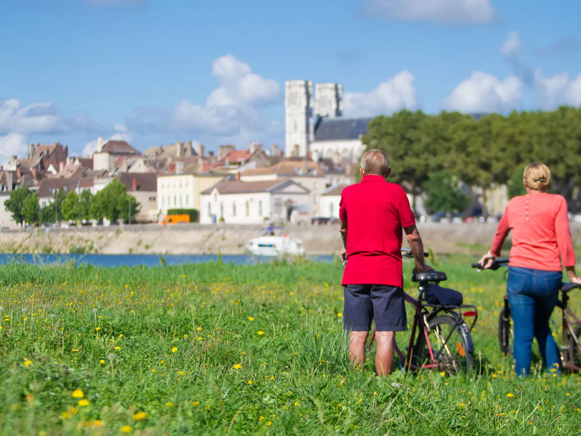 Rear view of an older man and woman with bicycles stood in a grassy field against a blue sky looking across a river at the buildings of Chalon-Sur-Saône in France