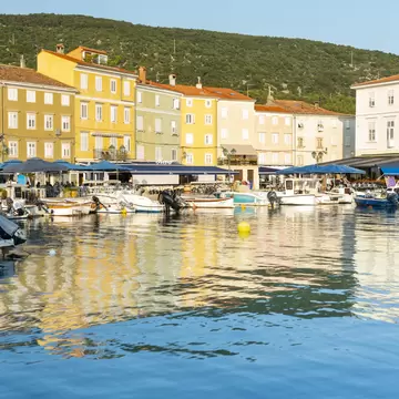 Yellow and cream houses with blue café umbrellas line Cres town harbour, reflected in calm water below a wooded hill.
