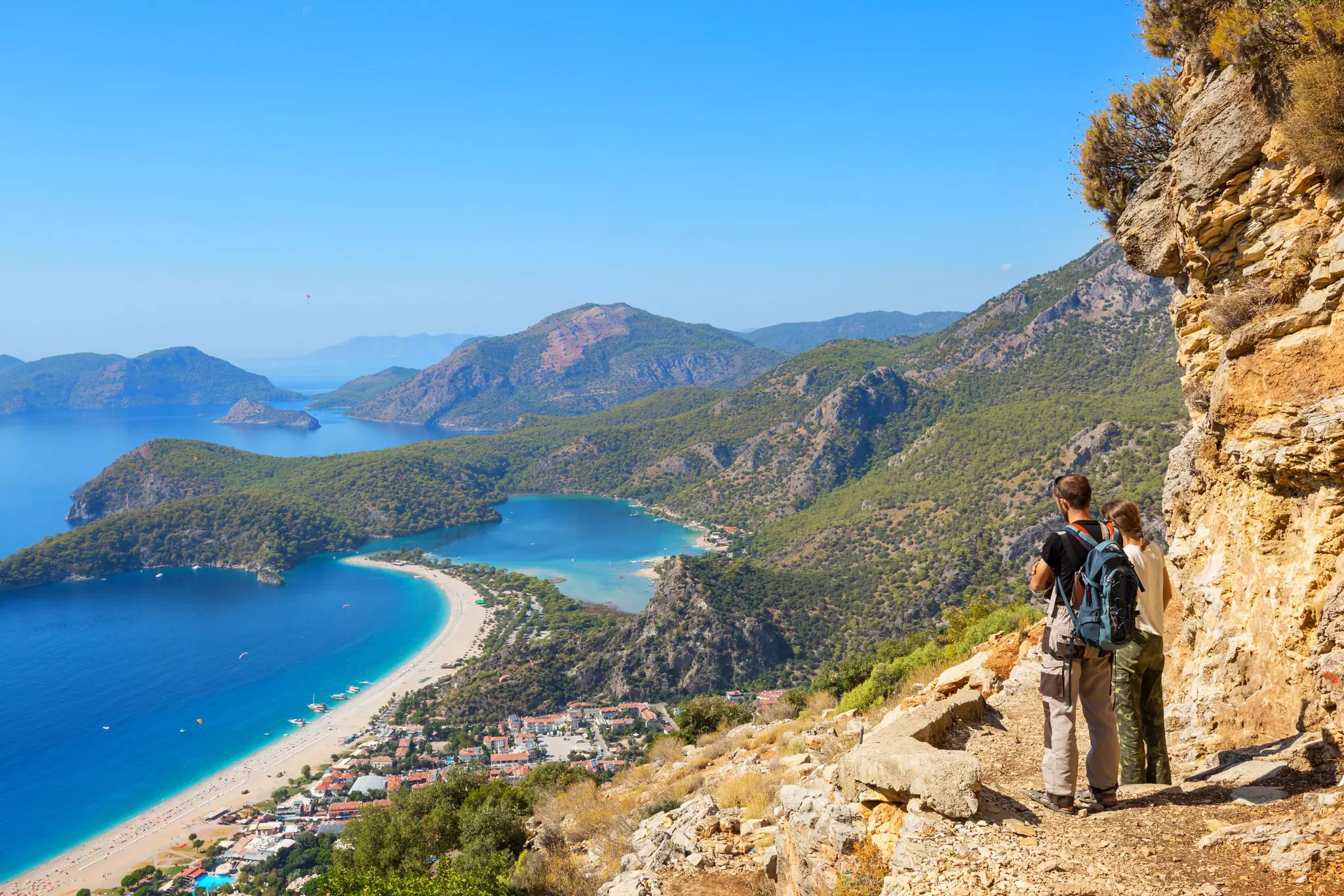 Two hikers on a rocky path in Türkiye look down at green hills and a beach by azure water.