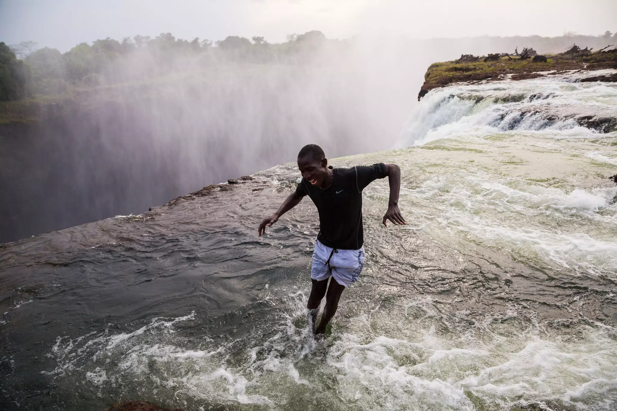 Two men in swim trunks enjoy Devil's Pool atop Victoria Falls, one standing close to the edge watching as the other jumps from a high point into the pool below with his back to the viewer