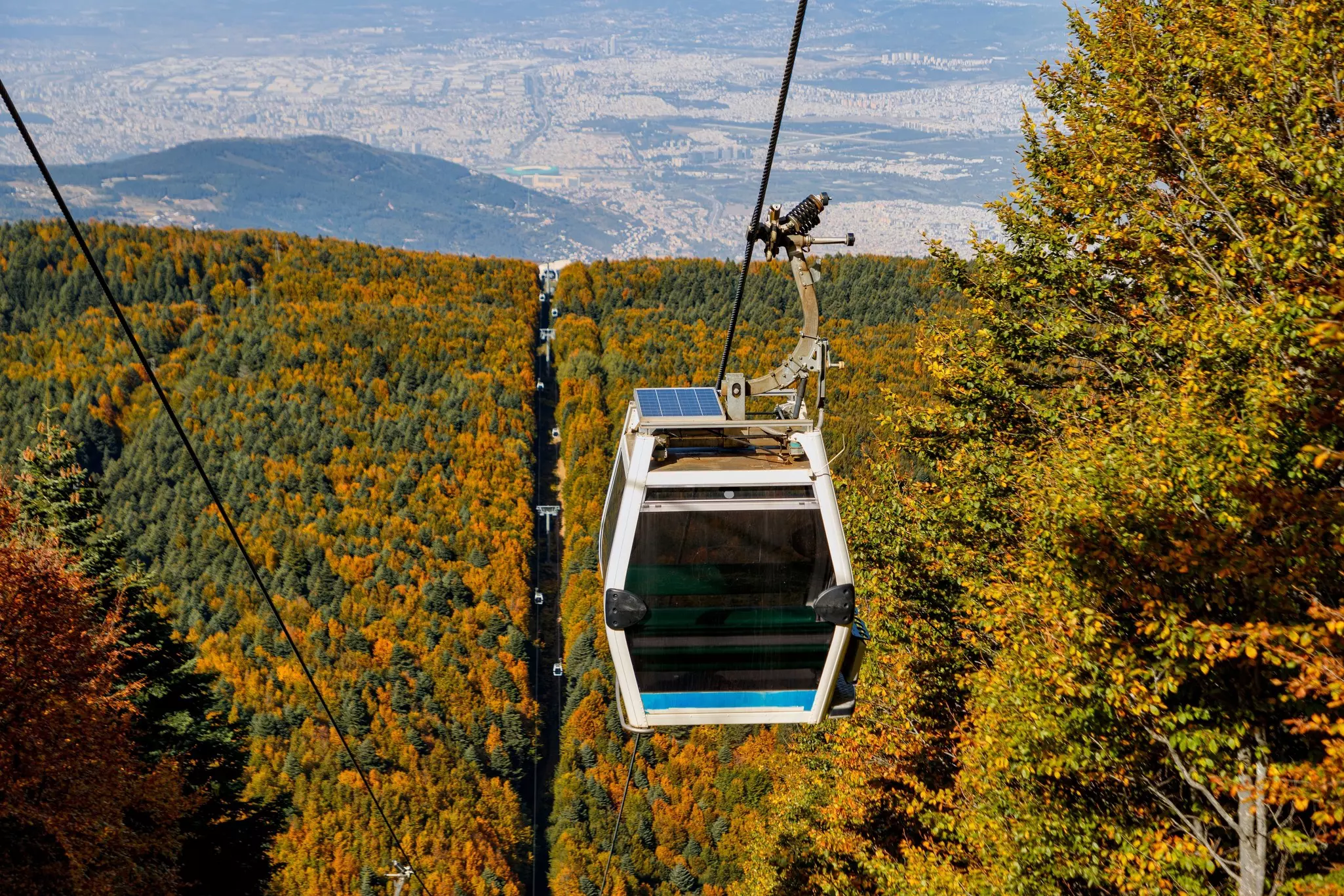 A gondola on a cable car ascends over a wooded hill colored in fall foliage. A huge city is seen below, in the distance.