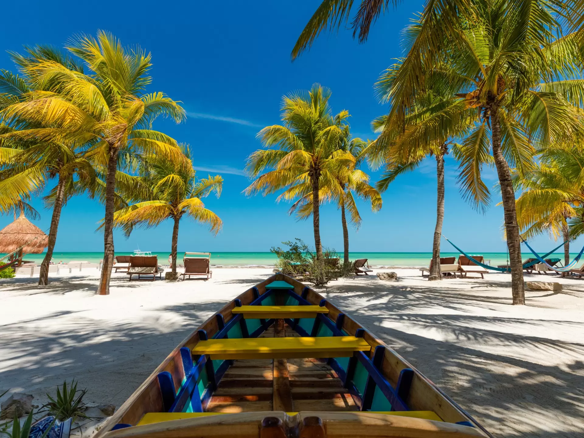 A brightly colored boat is moored in the sand, but facing the ocean and a collection of palm trees. There are a pair of lounge chairs to the left. 