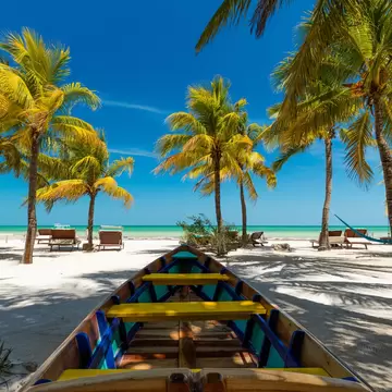 A brightly colored boat is moored in the sand, but facing the ocean and a collection of palm trees. There are a pair of lounge chairs to the left. 