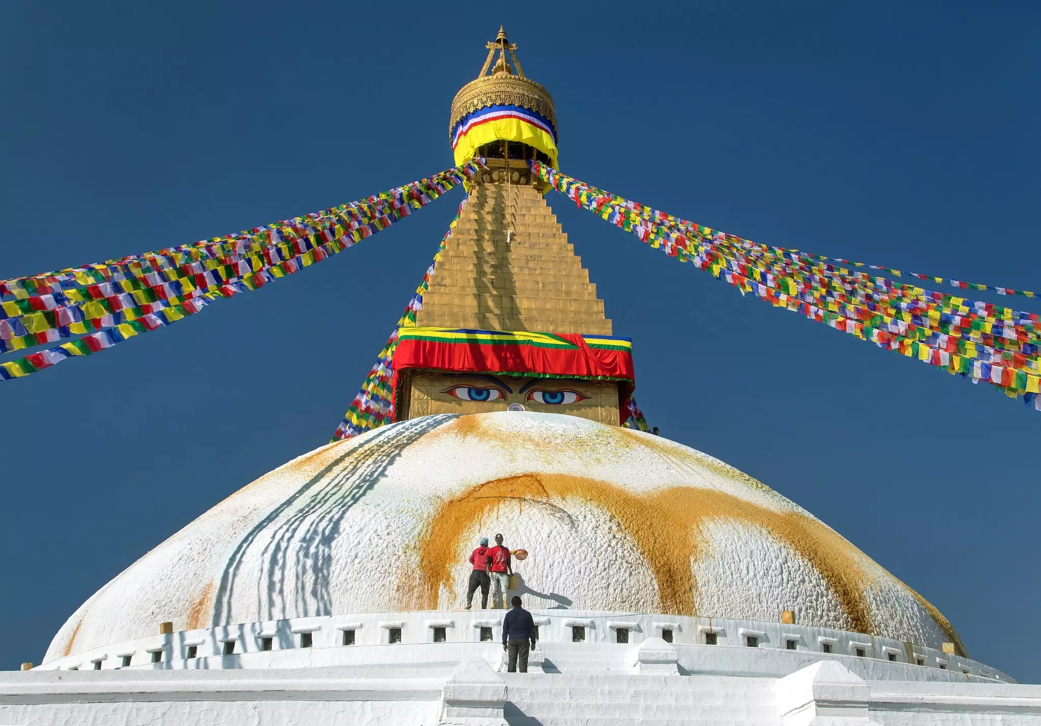 The painted white dome of a stupa in Nepal, with eyes painted on the tower and colorful flags extending from the top diagonally out of the frame.
