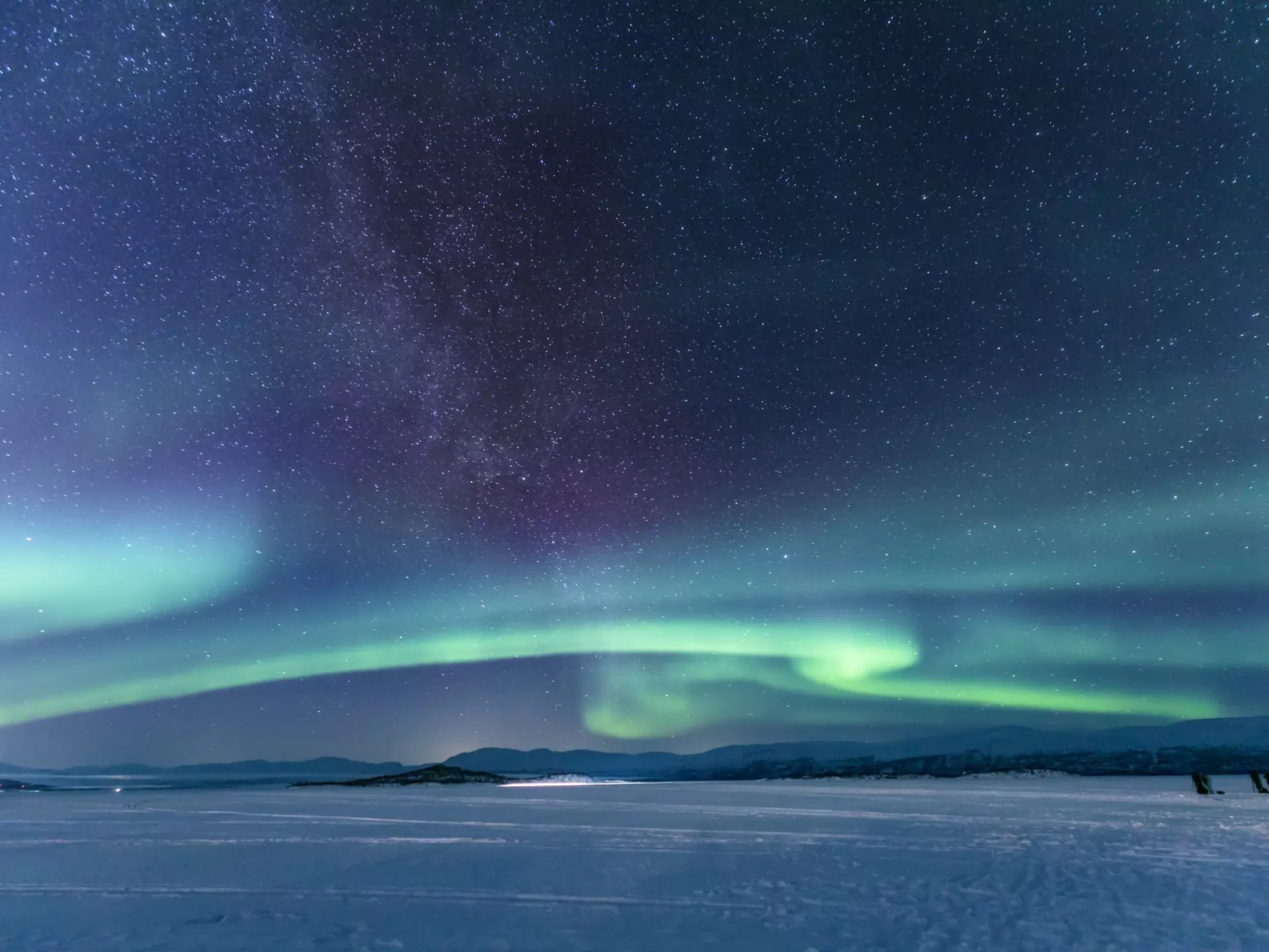 Green stripes of aurora borealis lights on a dark sky over a vast snowy field. 