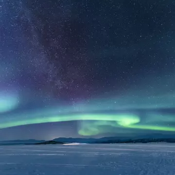 Green stripes of aurora borealis lights on a dark sky over a vast snowy field. 
