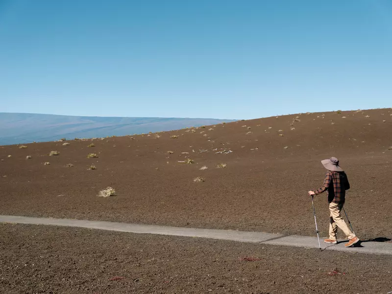 A solo hiker in a hat and with hiking poles walks along a narrow concrete path surrounded by dirt-covered hills on a sunny day.
