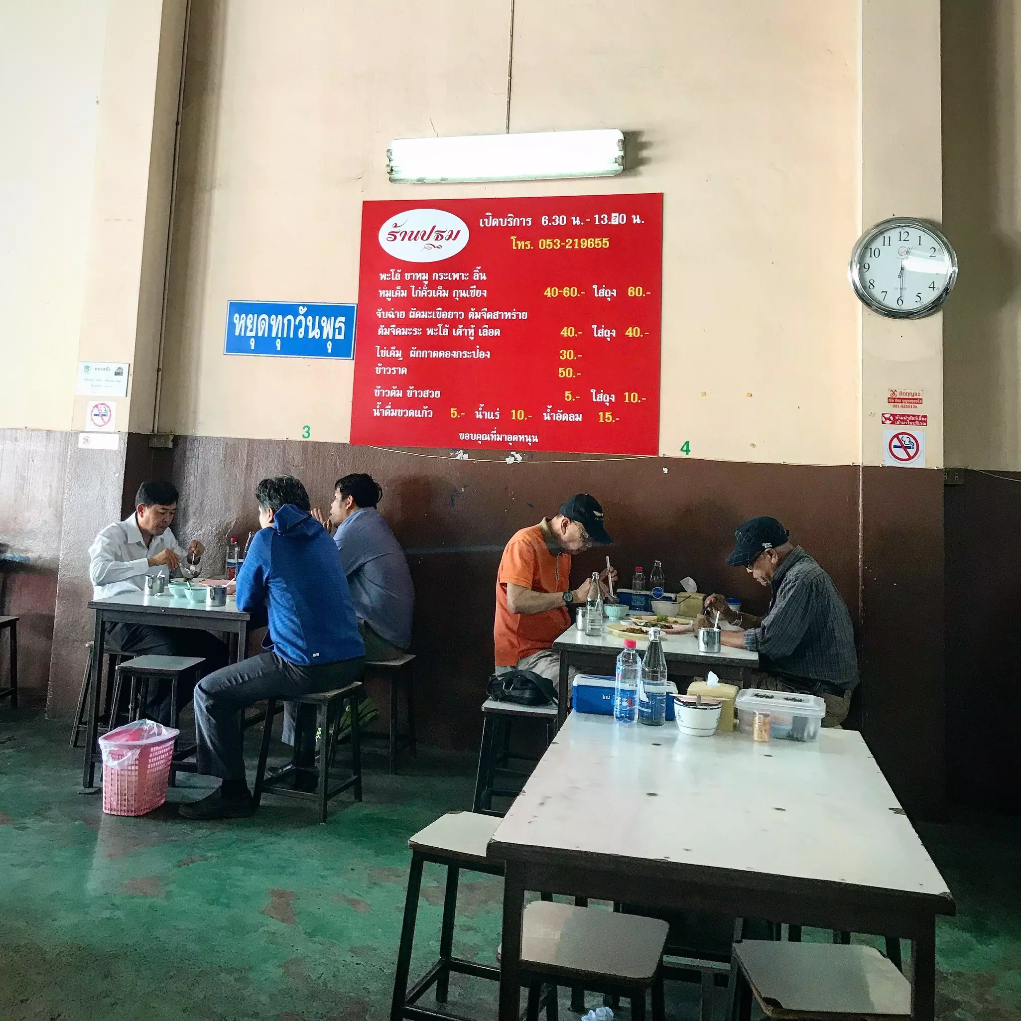 Tables of patrons at a simple cafe with a clock and two signs in Thai on its wall