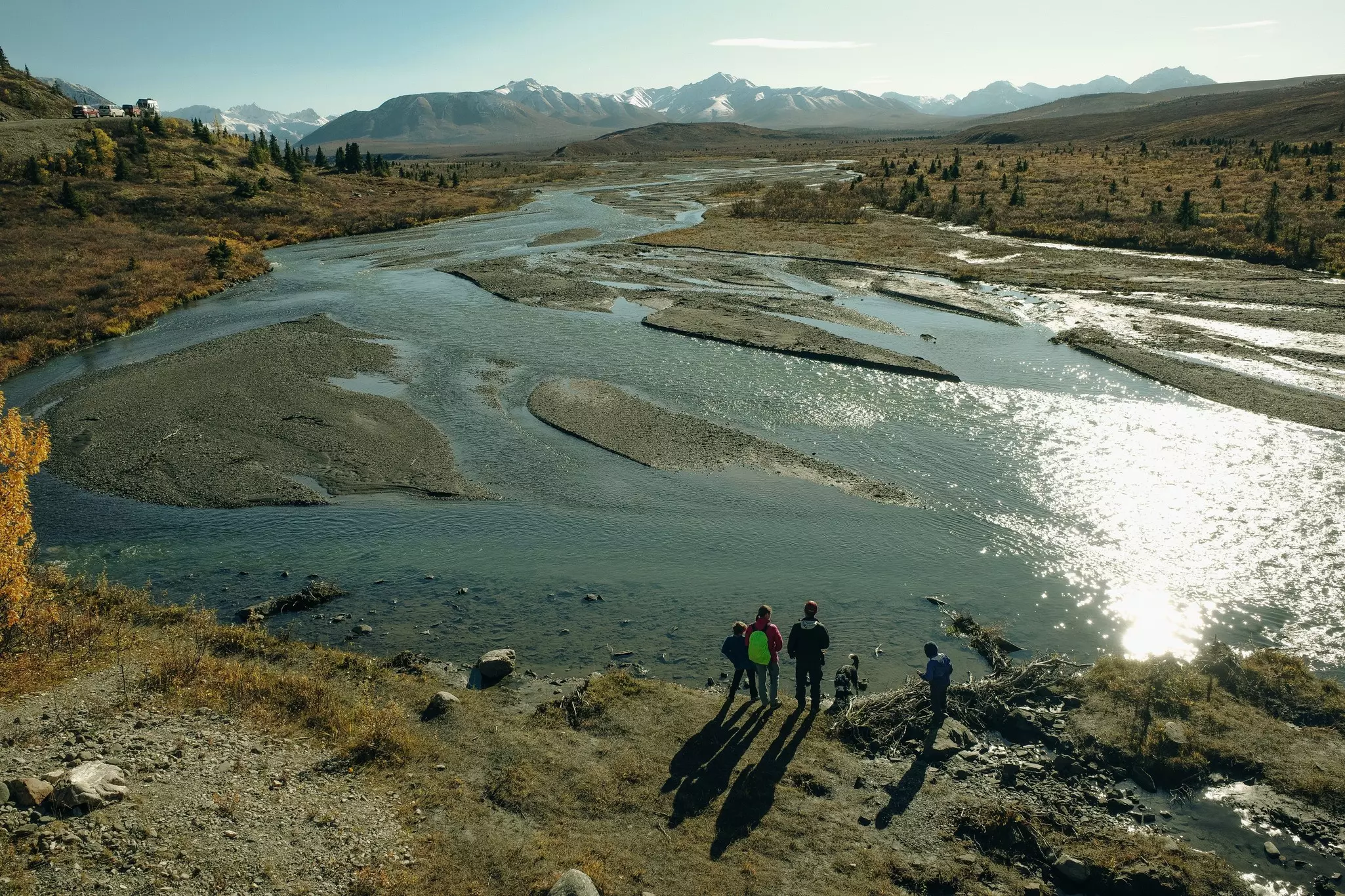 A family stands by the shores of a river. They're carrying backpacks and wearing hiking gear. Around them, are mountains and trees.