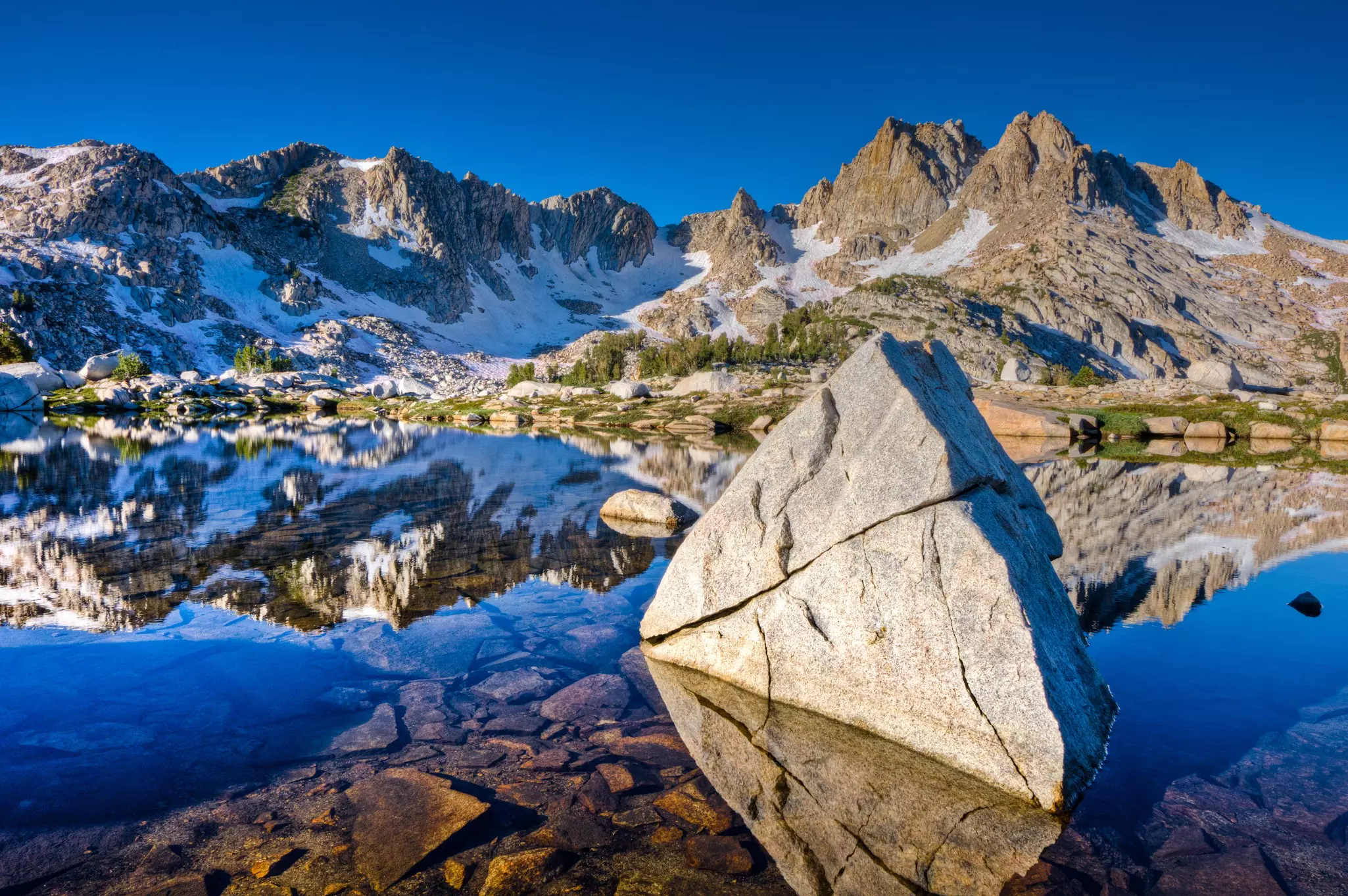 Rocks are reflected in the still waters of an alpine lake surrounded by mountains.