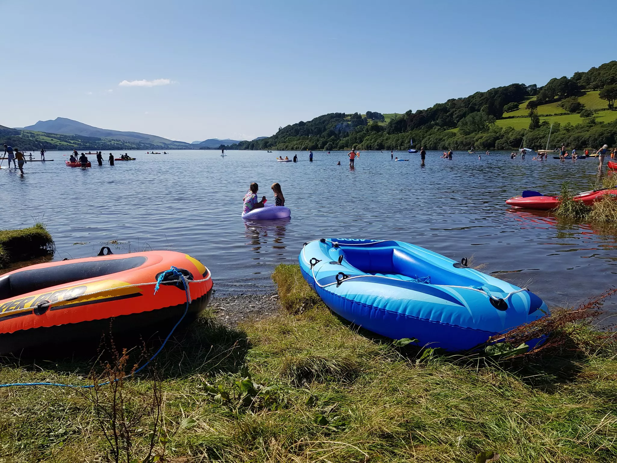 Inflatable boats on shore of Llyn Tegid as people participate in watersports on a sunny Bank Holiday in Wales.