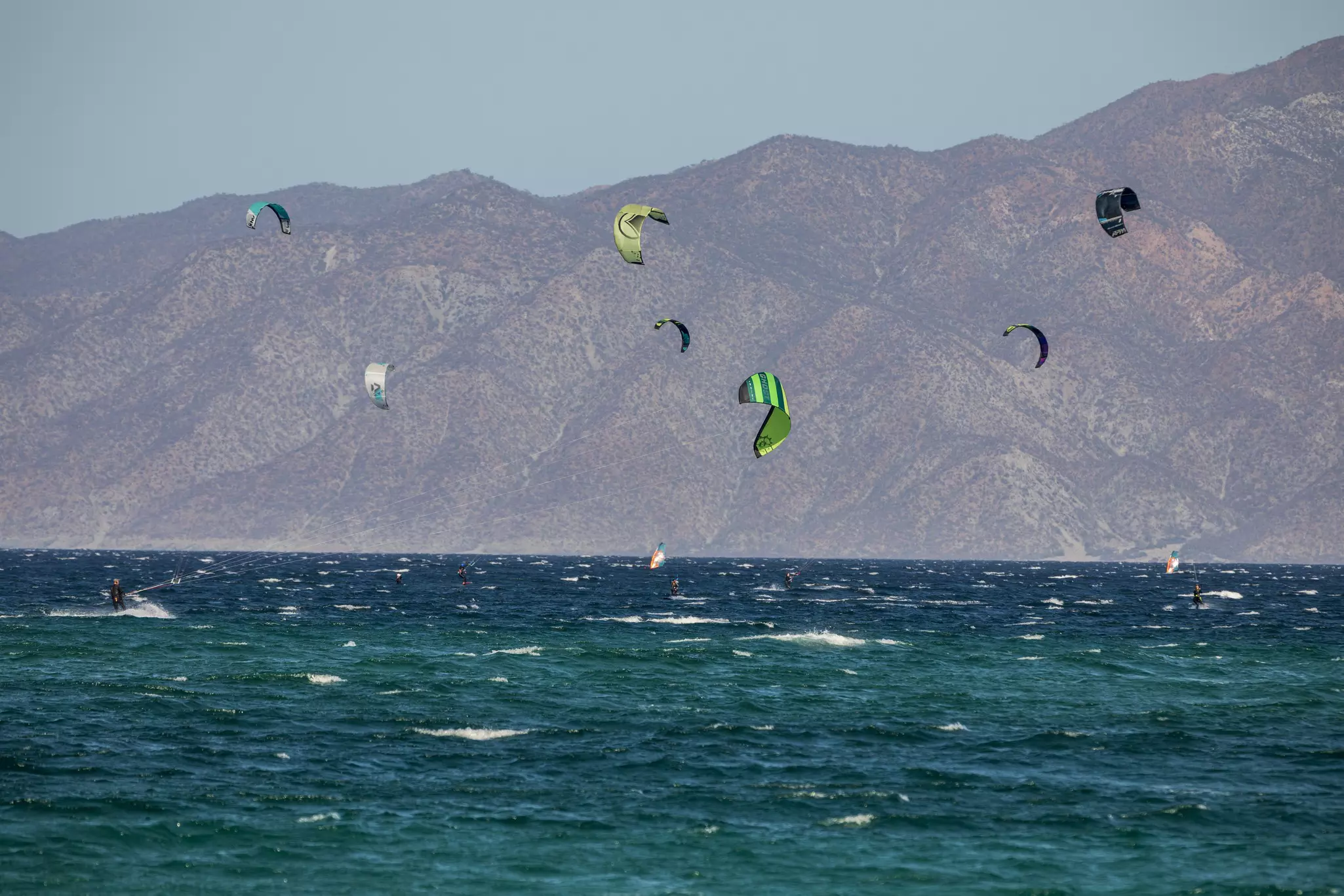 The sails of several kitesurfers above the white-capped sea, with tall mountains on the shore in the distance.