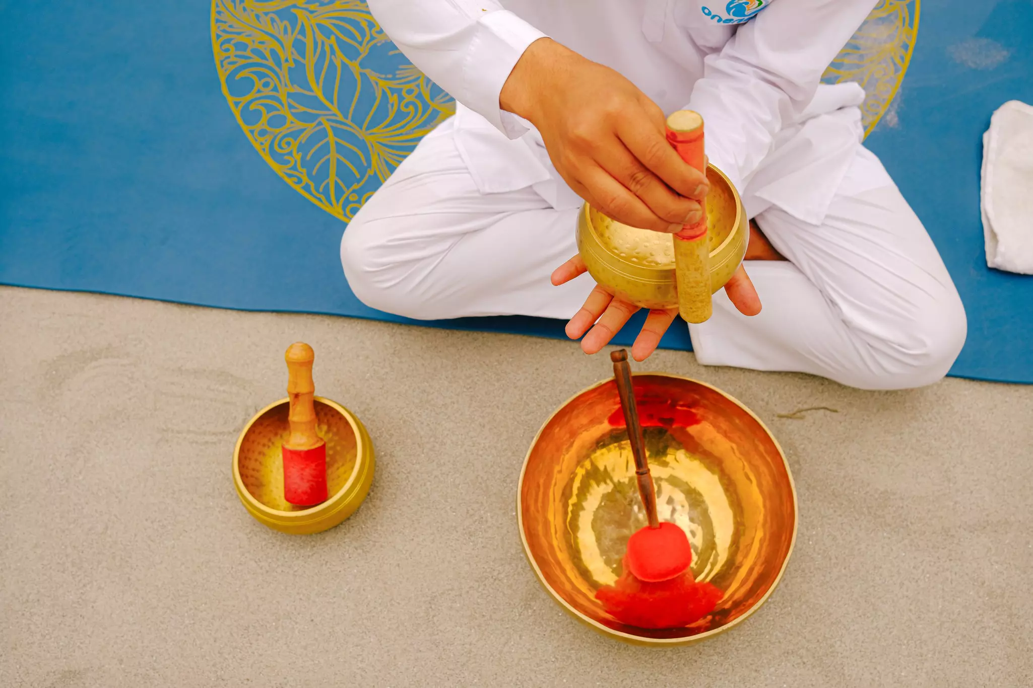 A person dressed in white and sat cross-legged on a mat runs a chime around a golden bowl.