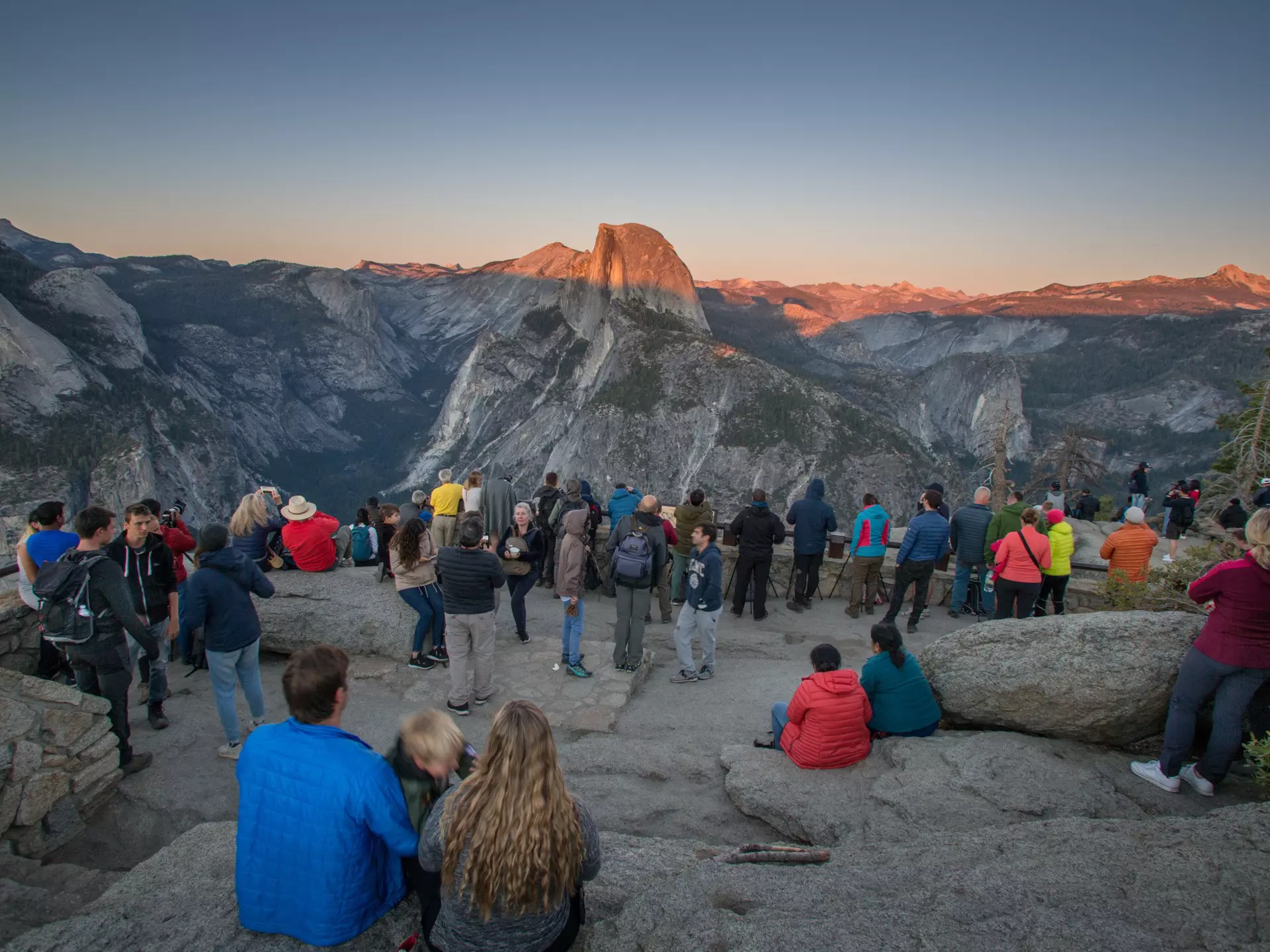People gather on a rocky ledge to watch the sun set over rock formations across a valley.