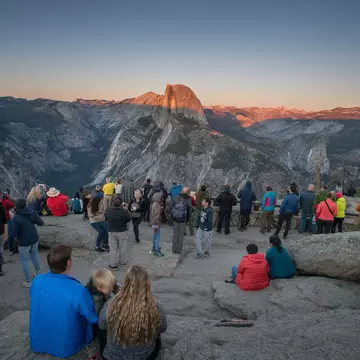 People gather on a rocky ledge to watch the sun set over rock formations across a valley.