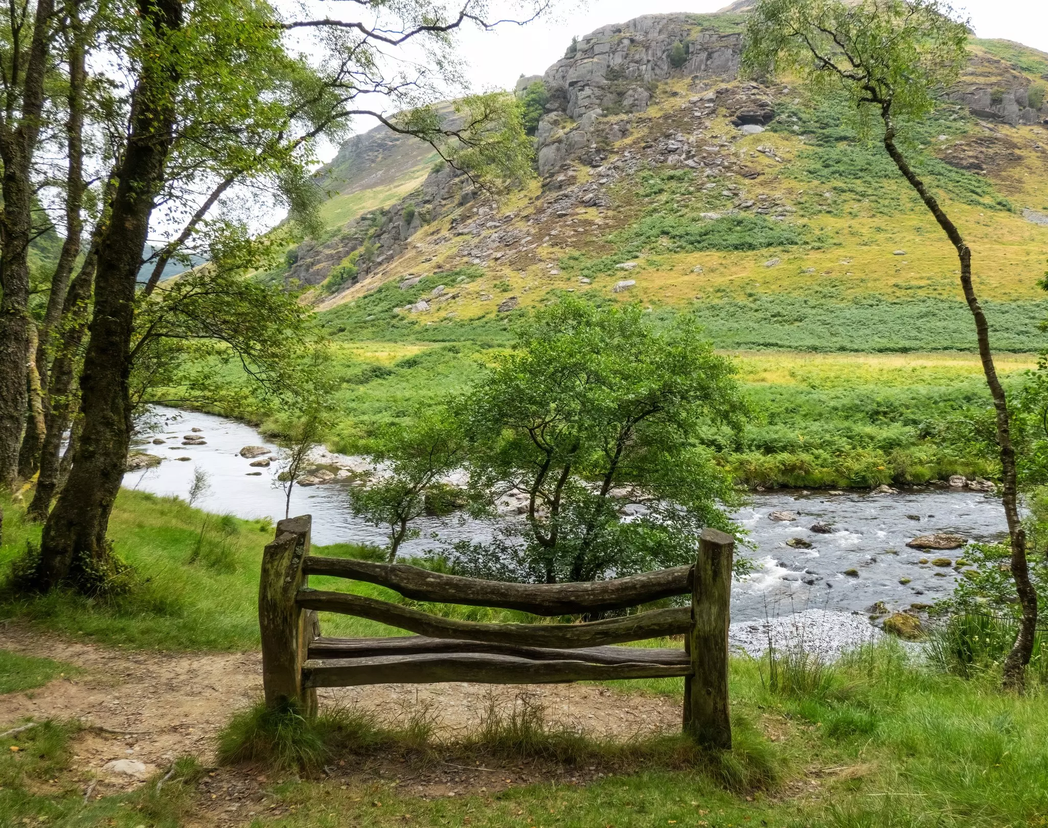 A wooden bench at the edge of a river looking towards a rocky outcrop