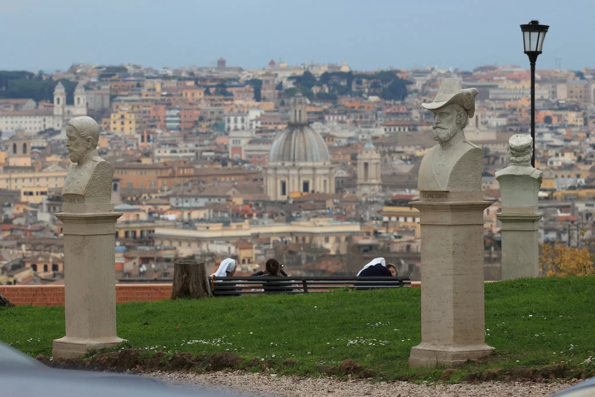 Nuns sitting on a bench atop Gianicolo Hill overlooking Rome, Italy, with statues in the foreground.