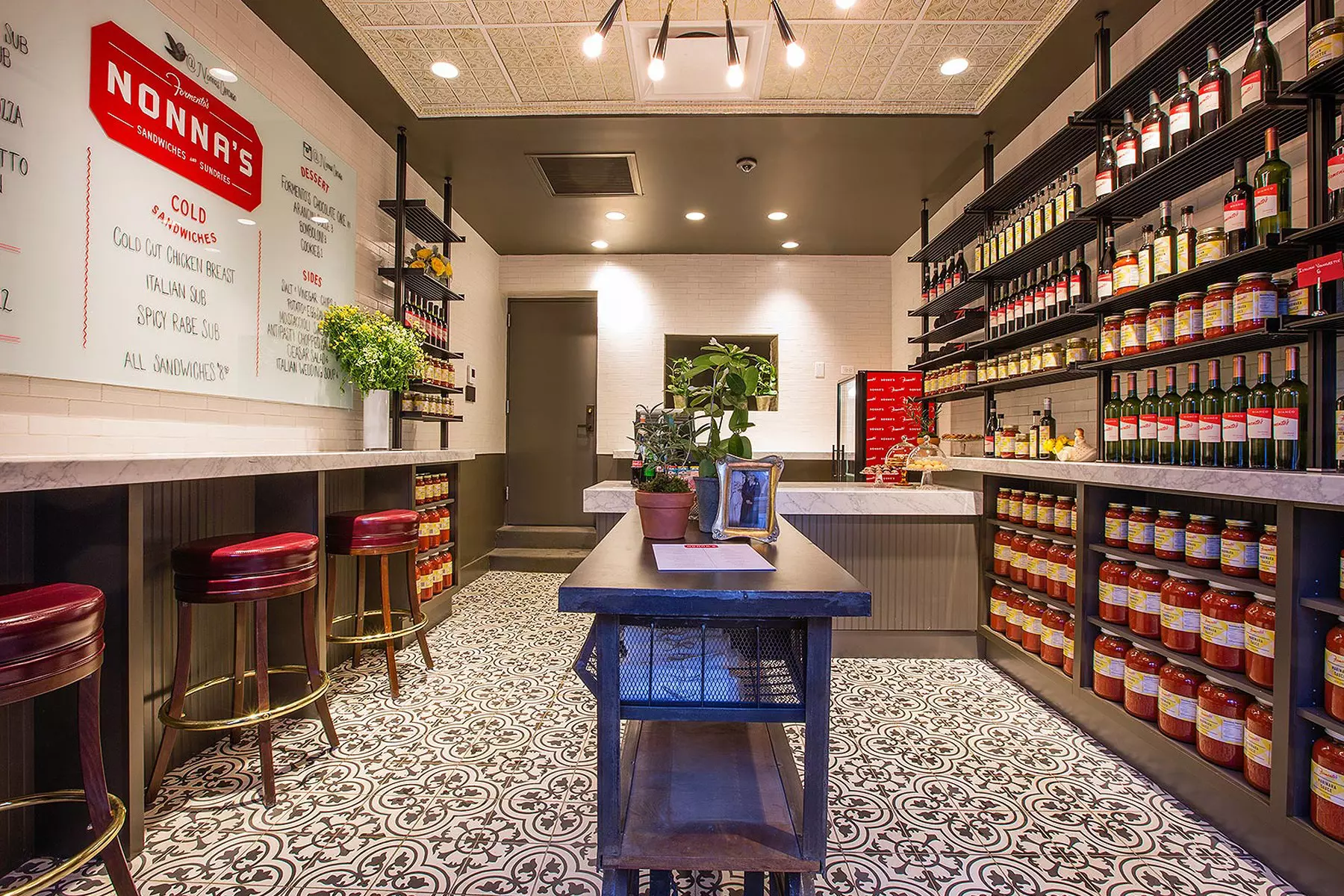 A restaurant interior with decorative black-and-white tiled flooring, jars and bottles in shelves along one wall, and a menu and counter with stools on the other wall.