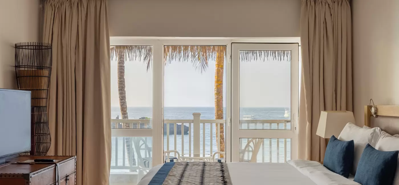 A white hotel bed with a blue striped blanket and blue pillows; a balcony with a view of the water is beyond the bed.