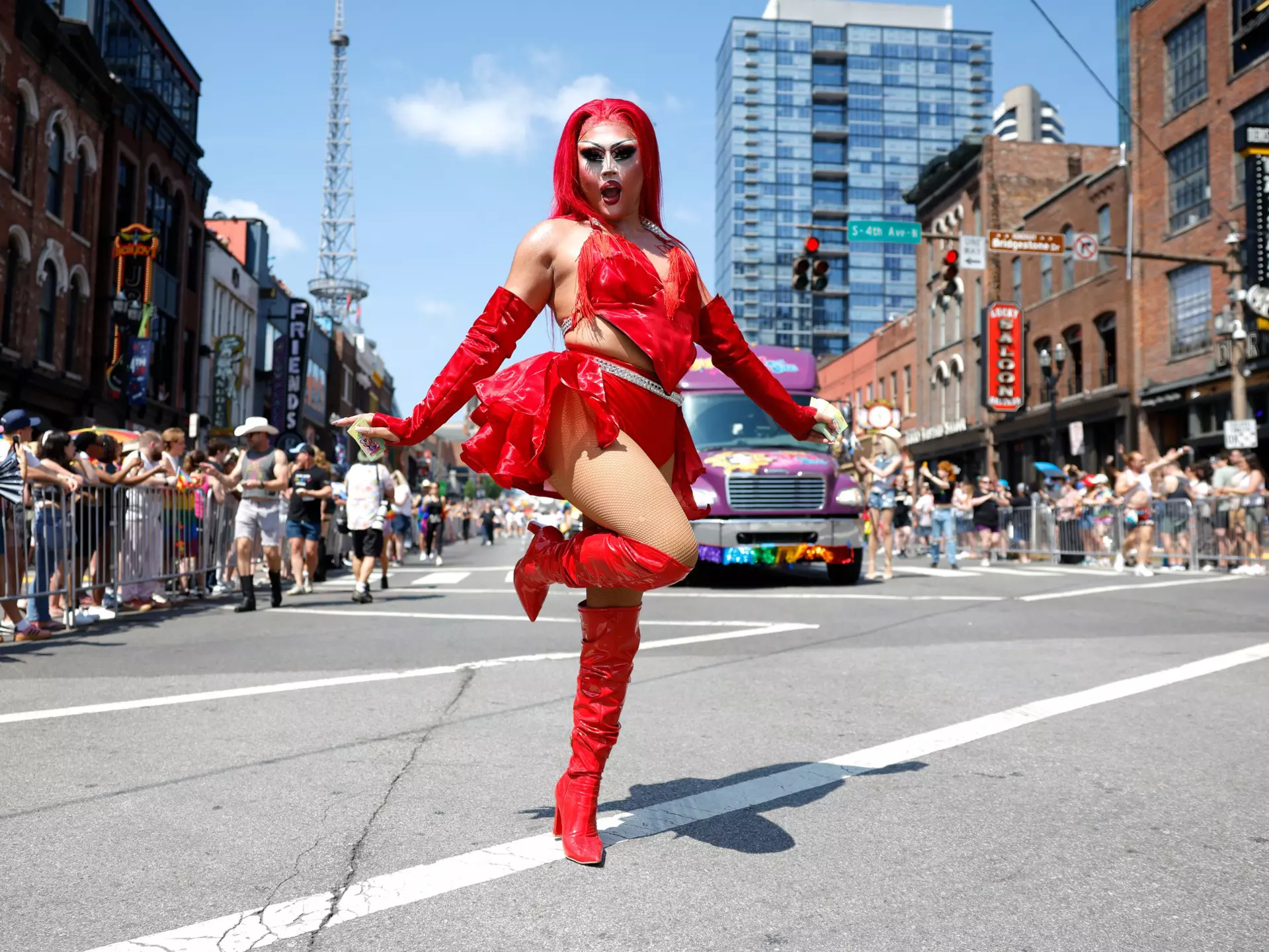 A drag performer in a red outfit and boots strikes a pose during a parade on a city street.