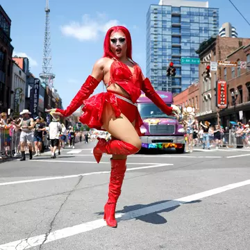 A drag performer in a red outfit and boots strikes a pose during a parade on a city street.