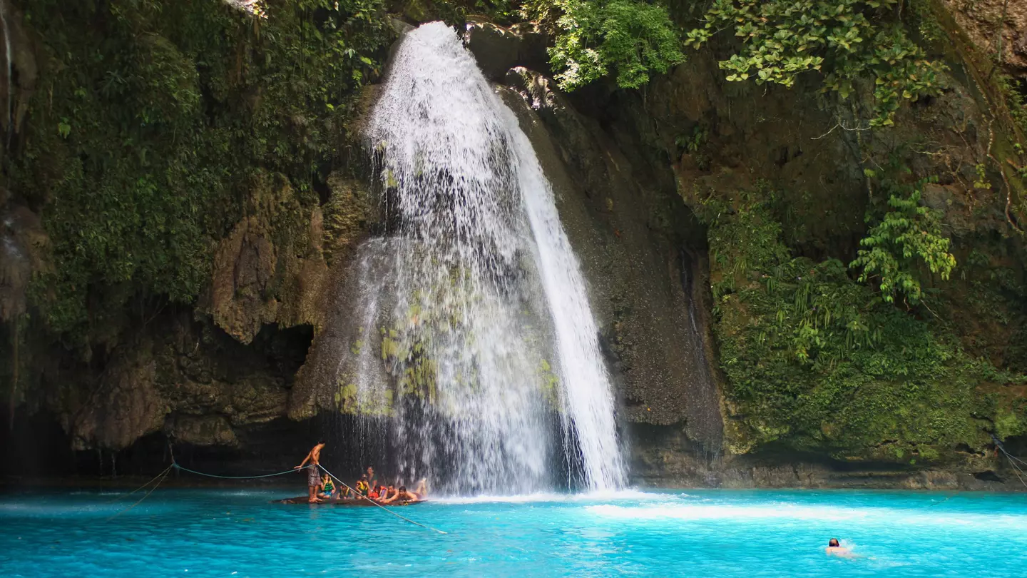 People swim in a blue lagoon at the base of a tall waterfall, which falls down a slope covered with greenery and rocks