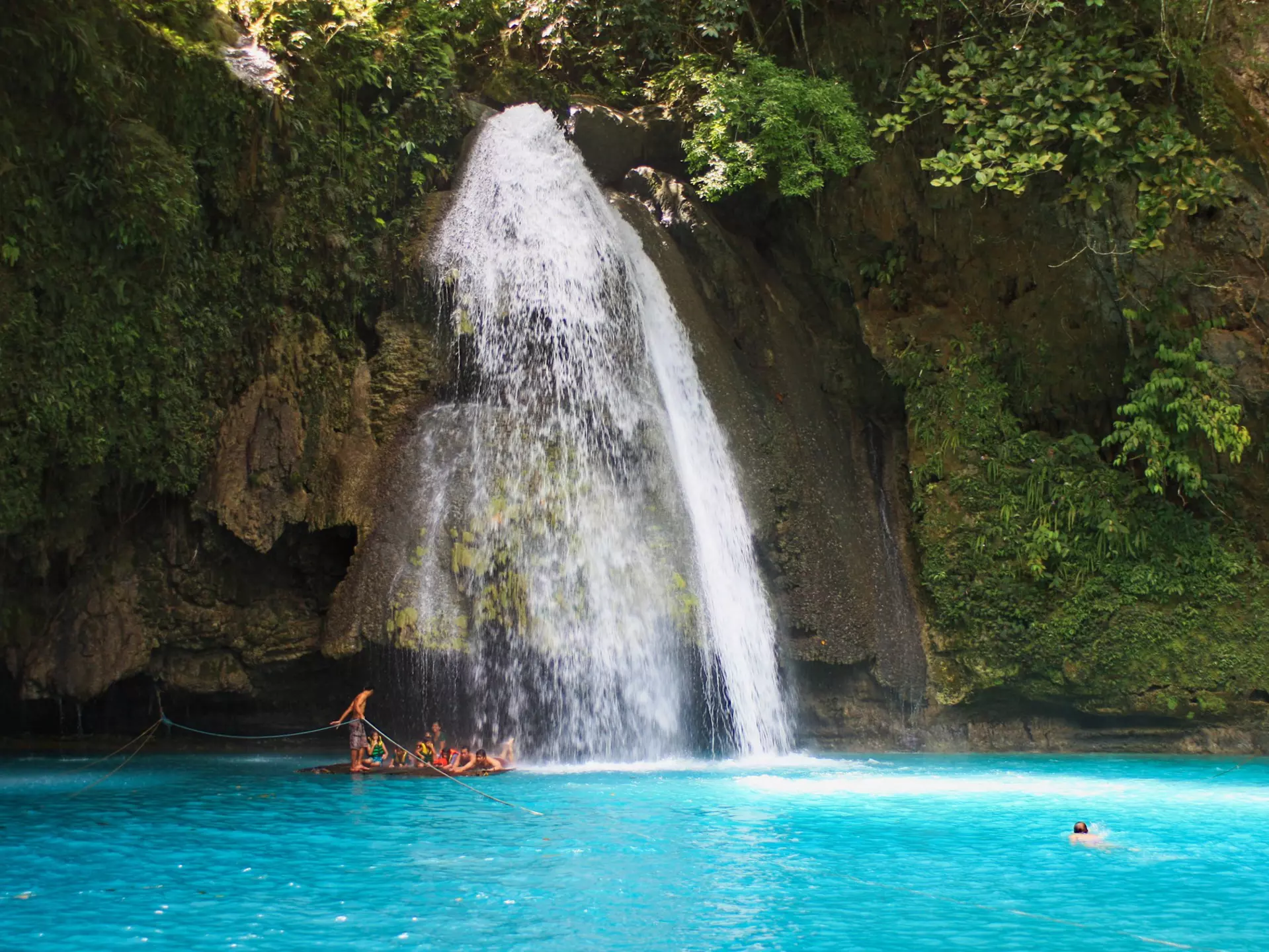 People swim in a blue lagoon at the base of a tall waterfall, which falls down a slope covered with greenery and rocks