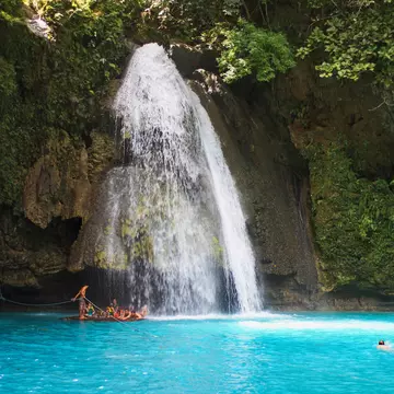 People swim in a blue lagoon at the base of a tall waterfall, which falls down a slope covered with greenery and rocks