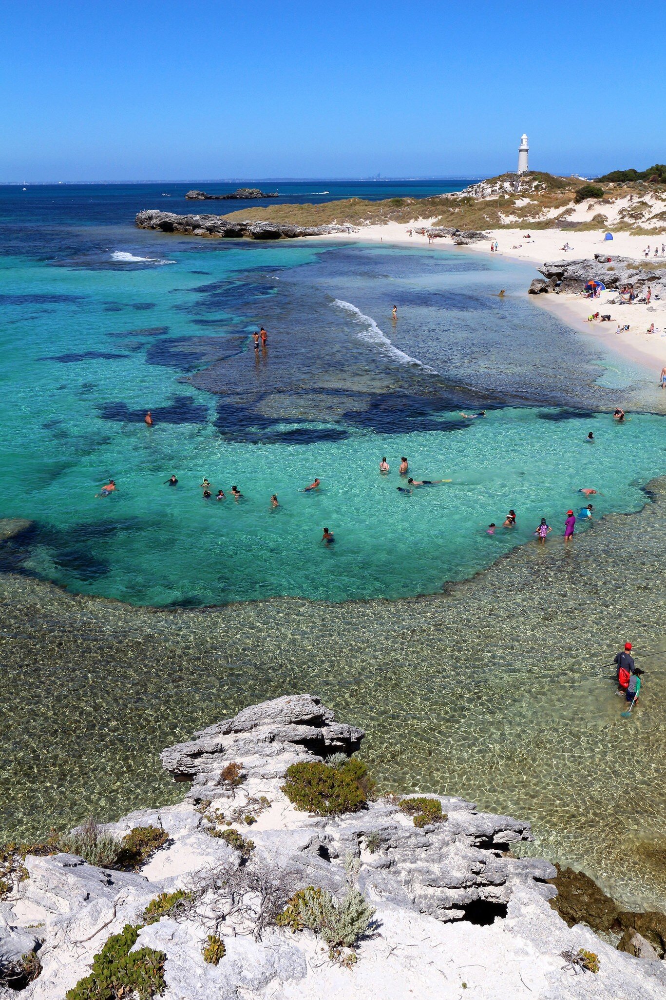 People swimming in turquoise water over dark reef beside a white-sand beach, a white lighthouse on the point, Rottnest Island