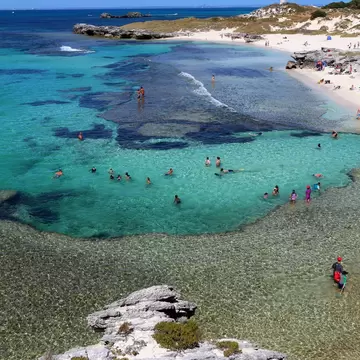 People swimming in turquoise water over dark reef beside a white-sand beach, a white lighthouse on the point, Rottnest Island