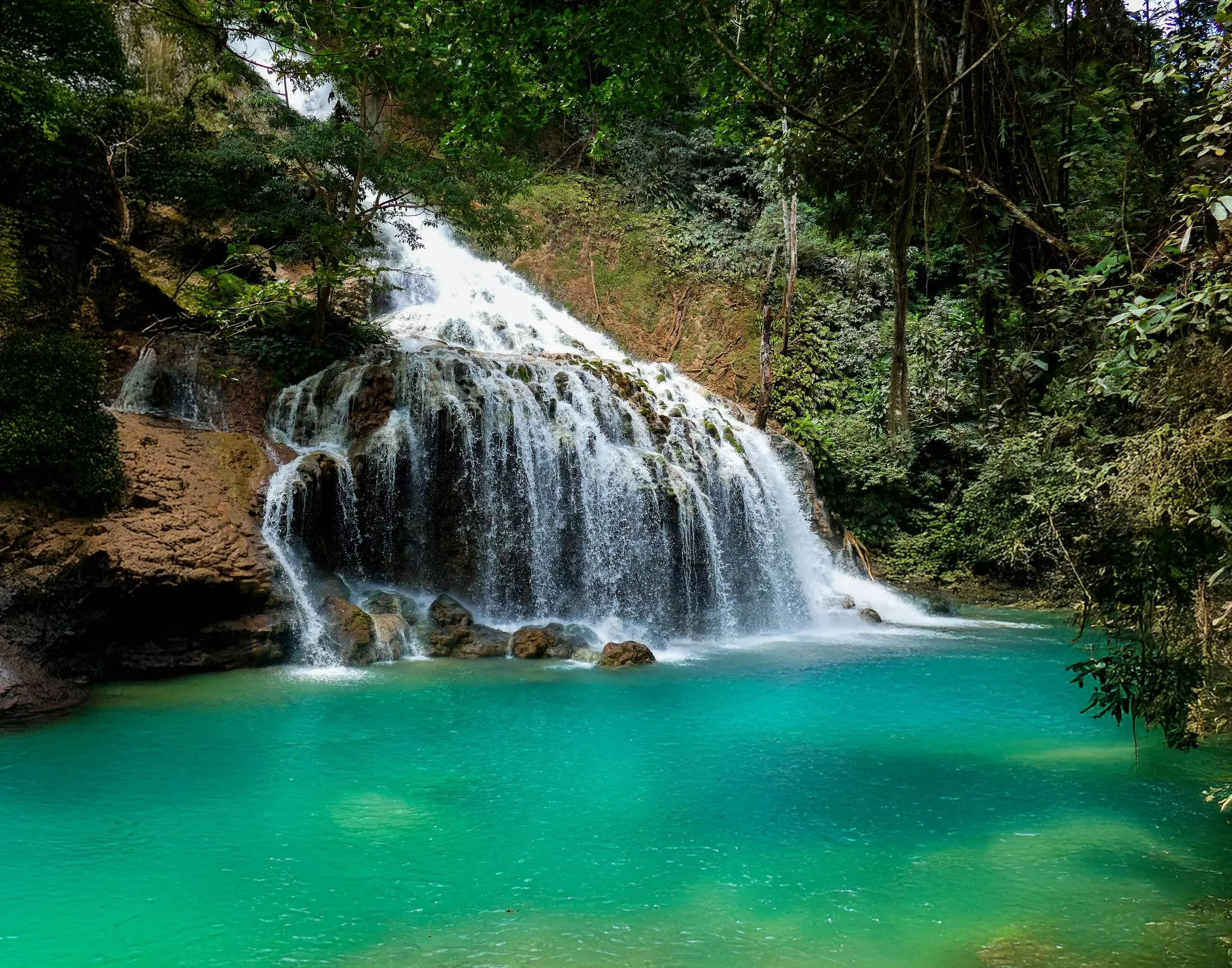 A view of a wide waterfall tumbling into a lagoon in a tropical forest.
