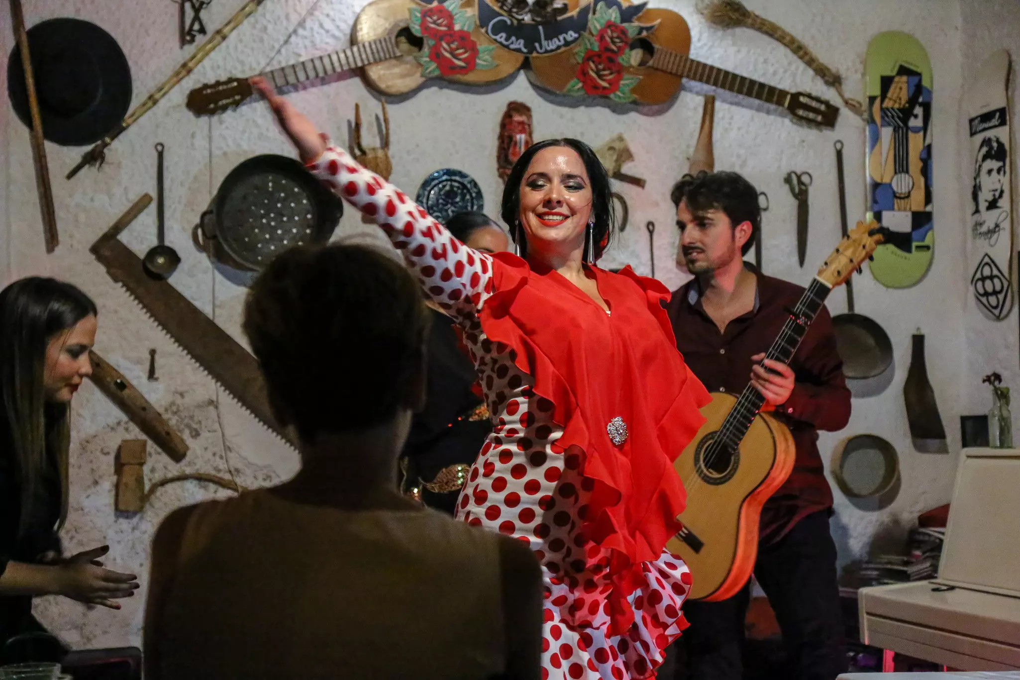 A flamenco dancer in a white dress with red polka dots wears a red shawl; musicians are playing behind her in front of a white wall with instruments and tools hanging on it.