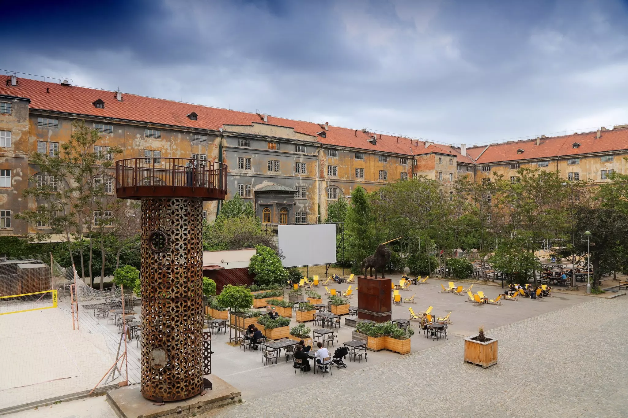 A large open-air courtyard surrounded by old warehouse buildings. People sit at tables and deck chairs.