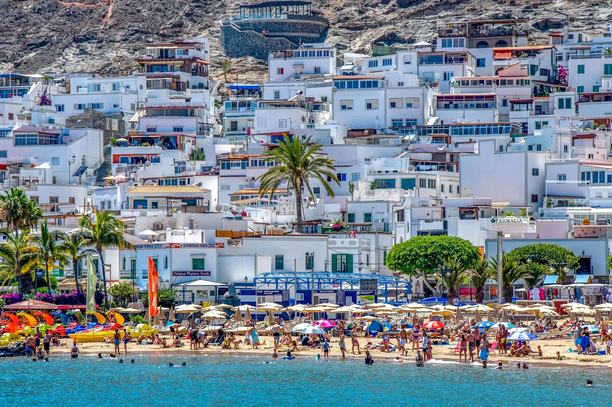 White buildings on a hillside are behind a beach crowded with people and umbrellas.