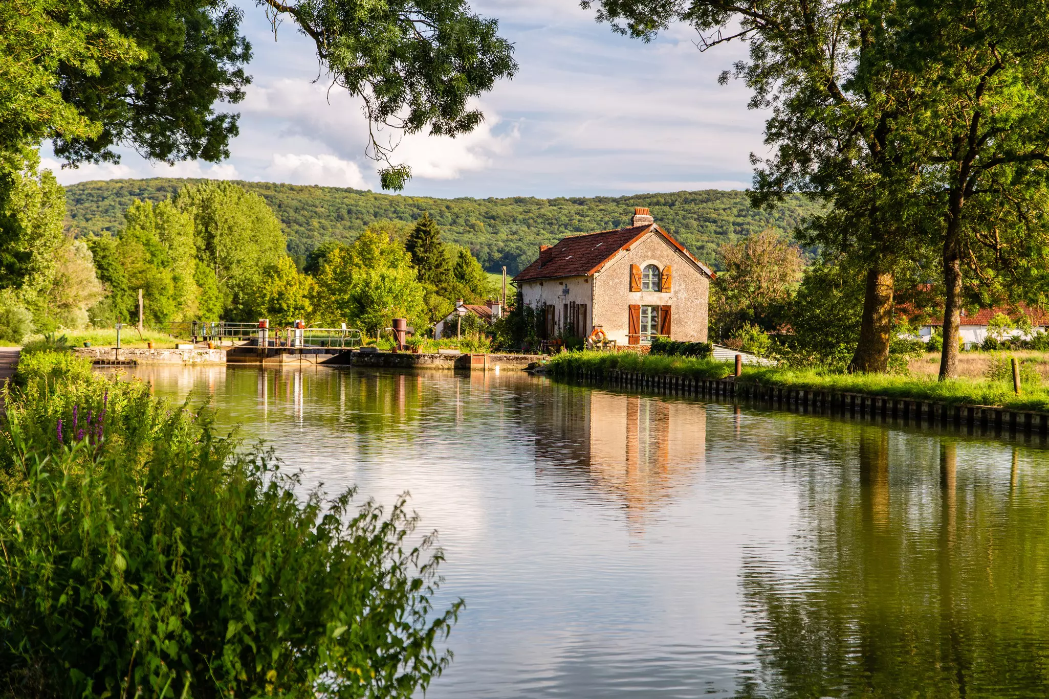 Stone house along a canal with a lock in the distance and trees bordering the wateron a sunny day. A forested hillside is in the far distance.